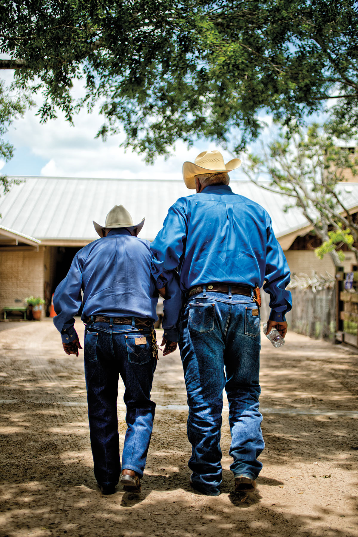 Two men, both wearing cowboy hats, blue shirts and blue jeans, walk along a dirt path